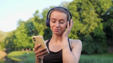 Koştuktan sonra şehir parkında durup müzik dinleyen gülümseyen pozitif bir kadının portresi. Spor yaptıktan sonra cep telefonu uygulaması kullanan güzel bir genç kadın. Tatmin olmuş kız