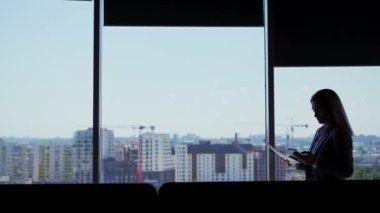 Silhouette of businesswoman using smartphone chatting online holding documents standing by window in office. Side view silhouette of executive woman walking along hall with panoramic windows