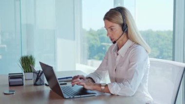 Confident female office worker wears headset having conference video calling looking at laptop screen. Businesswoman customer service manager speaking to webcam communicating in distance remote chat 