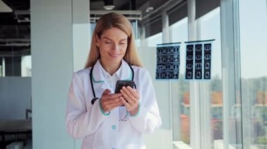 Smiling female doctor in medic coat and stethoscope using smartphone, standing near panoramic window. Focused physician consulting clients online via mobile healthcare application in office