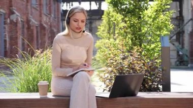 Pretty woman university student in headset learning online using laptop computer, taking notes, watching webinar virtual education remote class studying outdoor sitting outside college campus area