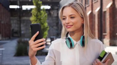 Close up of the young caucasian student woman having a video chat talking with friend standing in university campus outdoors. Charming beautiful happy girl talking online using mobile phone app