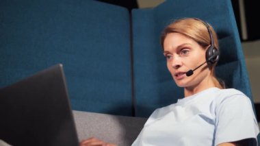 Confident middle aged pretty businesswoman wearing headset with mic sitting in coworking center enjoying video online conference call with foreign partners indoor