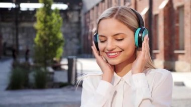 Beautiful blonde woman listening to music in wireless headphone using mobile smartphone new app in urban street take off modern headphones looking smiling in camera showing OK sign with thumb up