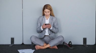 Stressed business woman without shoes sending message by mobile phone sitting near wall in office while meditate in lotus position. Young adult woman doing yoga exercise at work for stress relief
