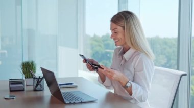 Glad happy business woman holding credit card using instant mobile payments at office workplace. Smiling beautiful girl lady customer shopper making online purchase on smart phone receiving cashback