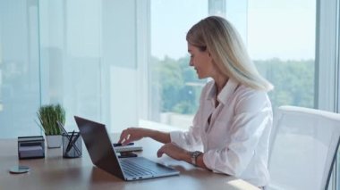 A woman is enthusiastically working on a laptop making notes in notebook in office. Professional blonde businesswoman sits at workplace desk near window panoramic. Happy employee working typing on pc
