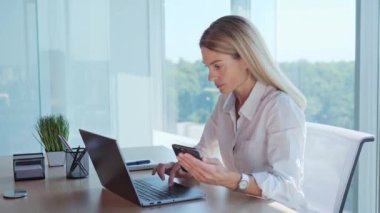 An office worker makes a calculation, a financial report, uses wireless Internet technologies, a phone and laptop. Businesswoman sit in office hold smartphone enters data into the computer pc indoor