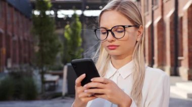 Beautiful blonde woman standing near authentic office building holding smartphone in hands. Pretty female freelancer using wireless network for modern device. Girl checking sending message outdoor