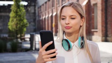 Young beautiful woman with headphones standing in town street having video online call on smartphone. Pretty stylish female student holding distance group video chat on mobile phone near academy
