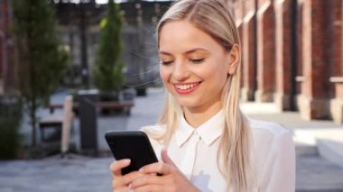 Caucasian smiling successful businesswoman in white shirt standing in urban street, taping on mobile phone communicates in social networks scrolling smartphone outdoor. Female freelancer relaxing