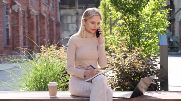 Молода Блондинка Робить Нотатки Блокноті Розмовляючи Мобільному Телефоні Зовні Красива — стокове відео
