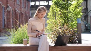 Charming young blonde attractive girl freelancer in casual modern clothes makes notes in notebook working with laptop outdoor. Female student doing homework remotely Education concept Uni campus area