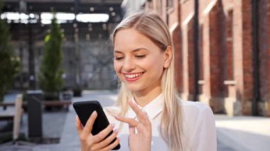 Positive young woman in white shirt uses mobile phone chatting with friend in social media. Joyful lady blond browsing internet date app, enjoys texting on smartphone outdoor. New modern technologies