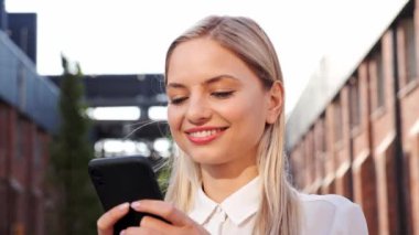Adorable Beautiful Woman checking Emails on Smartphone outdoors. Smiling Girl Female using Mobile Phone Chatting, Texting Message, Browsing Internet, Reading News . Successful Person. Smartphone App