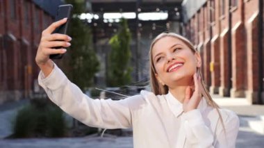 Closeup portrait of cheerful young blonde woman making selfie photos having fun video chat using smartphone. Amazed charming happy lady surprised looking at camera standing outdoors on city street
