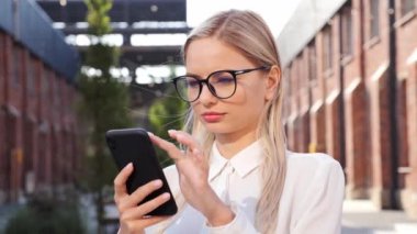 Confident serious freelancer in eyeglasses stands in business district uses phone scrolling tapping on smartphone new technology communication sunny day, social networks, female texting on gadget