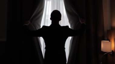A successful businessman in a formal suit opens the curtains in a hotel room during a business trip in the morning. Business man silhouetted shot from back. Confident male CEO top manager new day