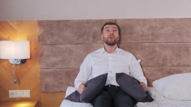 A businessman sits in formal clothes in a hotel bed and is not allowed to rest. A man puts pillows to his head because of noise isolation problems. A stressful day at work on a business trip