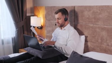 Young businessman customer service manager speaking to webcam laptop communicating in distance remote chat holding documents in hotel room. Male office worker wears headset conference video calling