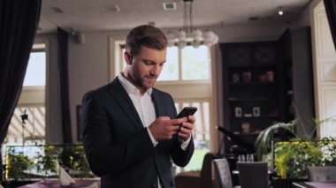 Positive businessman holding smartphone standing in restaurant. Middle aged manager ceo using cell phone mobile apps. Digital technology applications and solutions for business corporate development