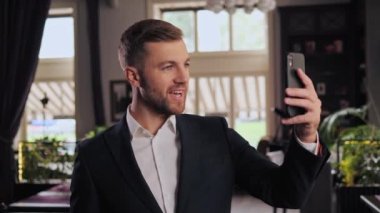 Portrait of young business man smiling and waving hand looking mobile phone camera having online video call chat in restaurant. Handsome guy having video conference. Leisure. New technology