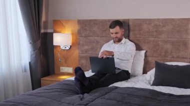 Handsome businessman open laptop computer sitting on a bed in luxury hotel, distance work concept. Man standing in hotel room. Wonderful Interior. Bos CEO Opens Laptop And Starts Working Indoor
