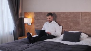 Lucky business man sitting on the bed working on laptop in hotel room celebrating success, amazing incredible news, gesturing hand, shouting yes winner. Financial stock sports betting crypto exchange
