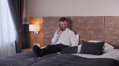 Smiling businessman calling by mobile phone in hotel room sitting on the bed. Happy business man talking on smartphone using laptop app distance working. Businesswoman call phone in a cozy room