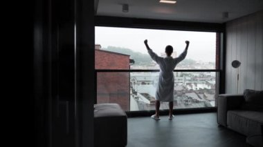 Funny Successful Businessman Dancing In a Bathrobe at Home or Hotel. A successful business man is dancing near panoramic window with a view of the old authentic city. Guy having fun going crazy
