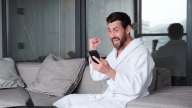 Excited happy businessman in bathrobe celebrating victory on phone in apartment, success winner guy looking at mobile phone in hotel. Closeup joyful man reading good news on phone Surprised lucky man