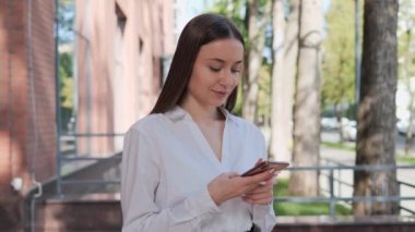 Güzel iş kadını dışarıda duruyor, mesajları kontrol ediyor, ofis merkezinin yakınındaki bir caddede akıllı telefon kullanıyor. Gülümseyen Güzel Kadın İşçi CEO 'su cep telefonunu dışarıda kullanıyor.