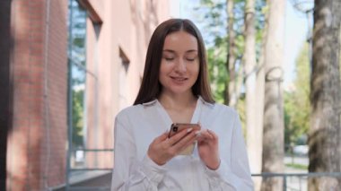 Beyaz tişörtlü sevimli genç iş kadını elinde akıllı telefonla dışarıda çalışan e-postalar yazıyor. Zeki bir kadın internette geziniyor. Yaz sabahı modern bir binanın yanında duruyor. İş adamları
