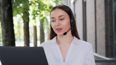 Gülümseyen genç iş kadını kablosuz kulaklık takıyor. Bilgisayardan video konferans görüşmesi yapıyor. Web kamerasından online sohbet, müşteri destek servisi, uzaktan eğitim kavramı.