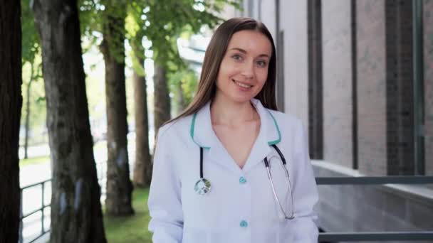 白い医療コートとカメラの屋外を見て聴診器を身に着けている幸せな若い美しい女性医師 病院の近くでポーズをとる女性医師の笑顔 積極的な開業医クローズアップ顔の肖像画 — ストック動画