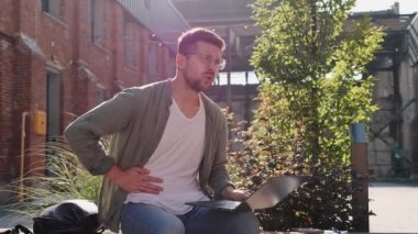 Kalitesiz yemek servisi. Akut mide ağrısı çeken serbest çalışan bir erkek işadamı, karaciğer ağrısı, ofisin yanındaki bankta oturuyor ve ağrılı karnına dokunuyor, dışarıda dizüstü bilgisayarla çalışan bir adam.