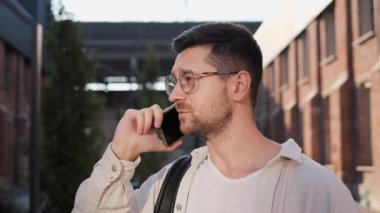 Sırt çantalı çekici bir adam akıllı telefonunu açık havada kullanıyor. Hoş ve ilginç bir görünüş. Yakışıklı bir adam cep telefonuyla dışarıyı arıyor. Erkek programcı konuşuyor.