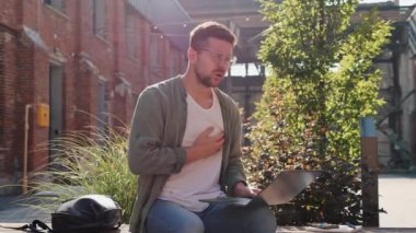 Kalp krizi, nefes darlığı, aritmi, günlük kıyafetli çekici bir adam dizüstü bilgisayarda çalışıyor, göğsüne dokunuyor, dışarıda kalp ağrısı çekiyor.