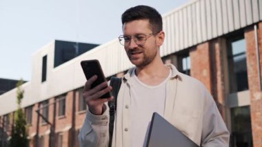 Gözlüklü yakışıklı sakallı adam telefon ekranına bakıyor ve caddede dikilirken gülümsüyor. Sosyal medyaya, internete, haberlere bakan yakışıklı bir adam.