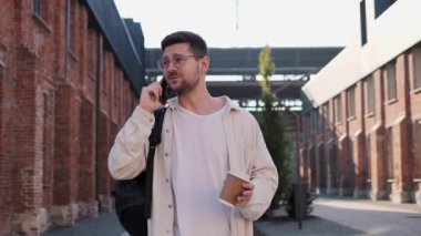 Şehirde şık giyinmiş, açık havada telefonla konuşan çekici bir adamın portresi. Gözlüklü adam elinde sıcak içecekle şehir caddesinde yürürken akıllı telefondan konuşuyor.