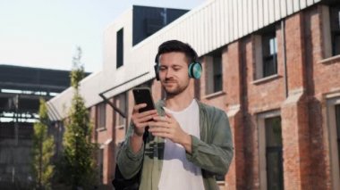 Çekici, şık, yakışıklı, kulaklıklı, akıllı telefondan müzik dinleyen bir adam. Mobil uygulamadan şarkı seçen bir adam. Dışarıda cep telefonu tutan, günlük kıyafetli neşeli yakışıklı bir adam.