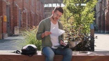 Genç kendine güvenen erkek üniversite öğrencisi dizüstü bilgisayarı kullanarak online belge okumayı öğreniyor sanal eğitim uzaktan eğitim semineri veriyor açık hava üniversitesi kampüs alanını inceliyor.