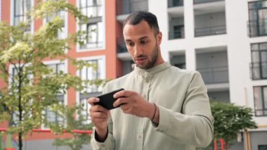 Afgan Amerikalı erkek kentsel iş adamı cep telefonu uygulamasını kullanarak çevrimiçi video oyunları oynuyor. Açık havada akıllı telefon kullanarak keyfine bakıyor.