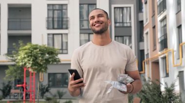 Elinde para sallayan yakışıklı Afrikalı Amerikalı adam, elinde cep telefonuyla para yatırarak para kazanmanın başarısını kutluyor. Mutlu adam dışarıda zaferi hissediyor.
