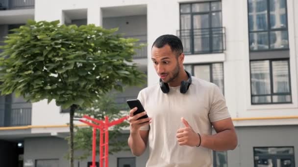 快乐积极的非洲裔美国人的肖像 使用智能手机 庆祝胜利 快乐地使用手机微笑暗色瘦身男性创业者 戴黑耳机的家伙 — 图库视频影像