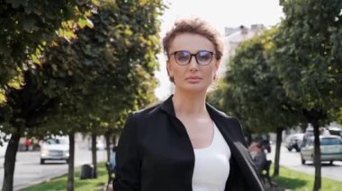 Açık havada duran gözlüklü, kendine güvenen güzel bir genç kadın portresi. Rüzgarlı havaya bakıyor. Ciddi iş kadını dışarıda poz veriyor, şehir arka planı, ağaçlar...