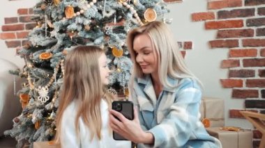 Güzel, güzel bir kız ve mavi gözlü bir anne akıllı telefon kullanarak arkadaşlarıyla konuşuyor. İnternetteki video konferans uygulaması ile Mutlu Noeller diliyor. Modern teknoloji Yeni Yıl 2023 Tebrikler