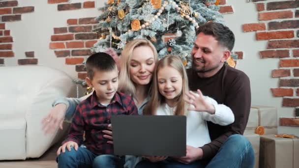 クリスマスツリーの近くに座っている幸せな興奮した家族は クリスマスの前夜の会議で家族や友人に挨拶するためのオンラインビデオ通話を行うラップトップを保持しています4人の家族 — ストック動画
