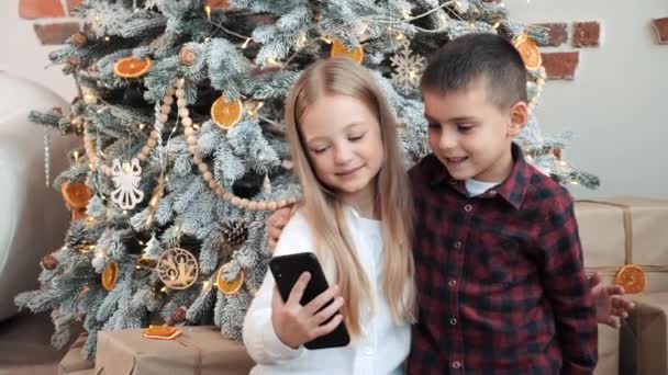 美しいブロンドの妹は 祖母とのオンラインビデオ通話中に弟を抱擁し クリスマスツリーの近くの自宅で空気のキスを送信します かわいい子供たちは彼女の両親を呼び出しますメリークリスマス — ストック動画