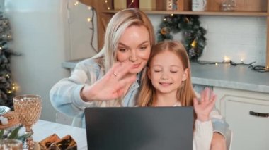 Mutlu bir anne ve kızı mutfakta sandalyede oturmuş görüntülü konuşma yapıyorlar. Oda bir Noel ağacı ve çelenklerle süslenmiş. Anne ve küçük kız el sallıyor.